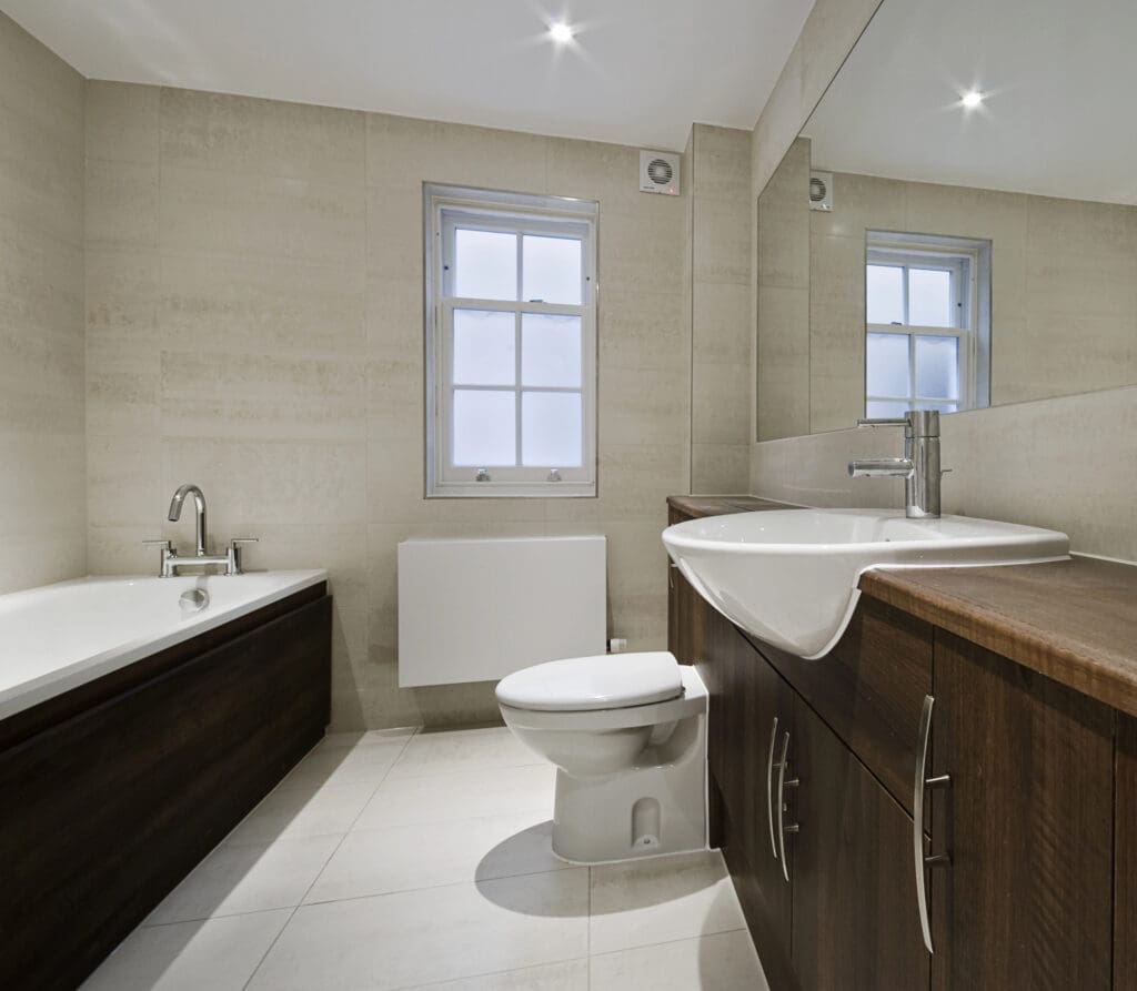 Modern bathroom with a large mirror, white sink set in dark wood cabinet, toilet, bathtub with matching dark wood panel, tiled floor and walls, and a window with frosted glass—ideal inspiration for your next bathroom renovation.