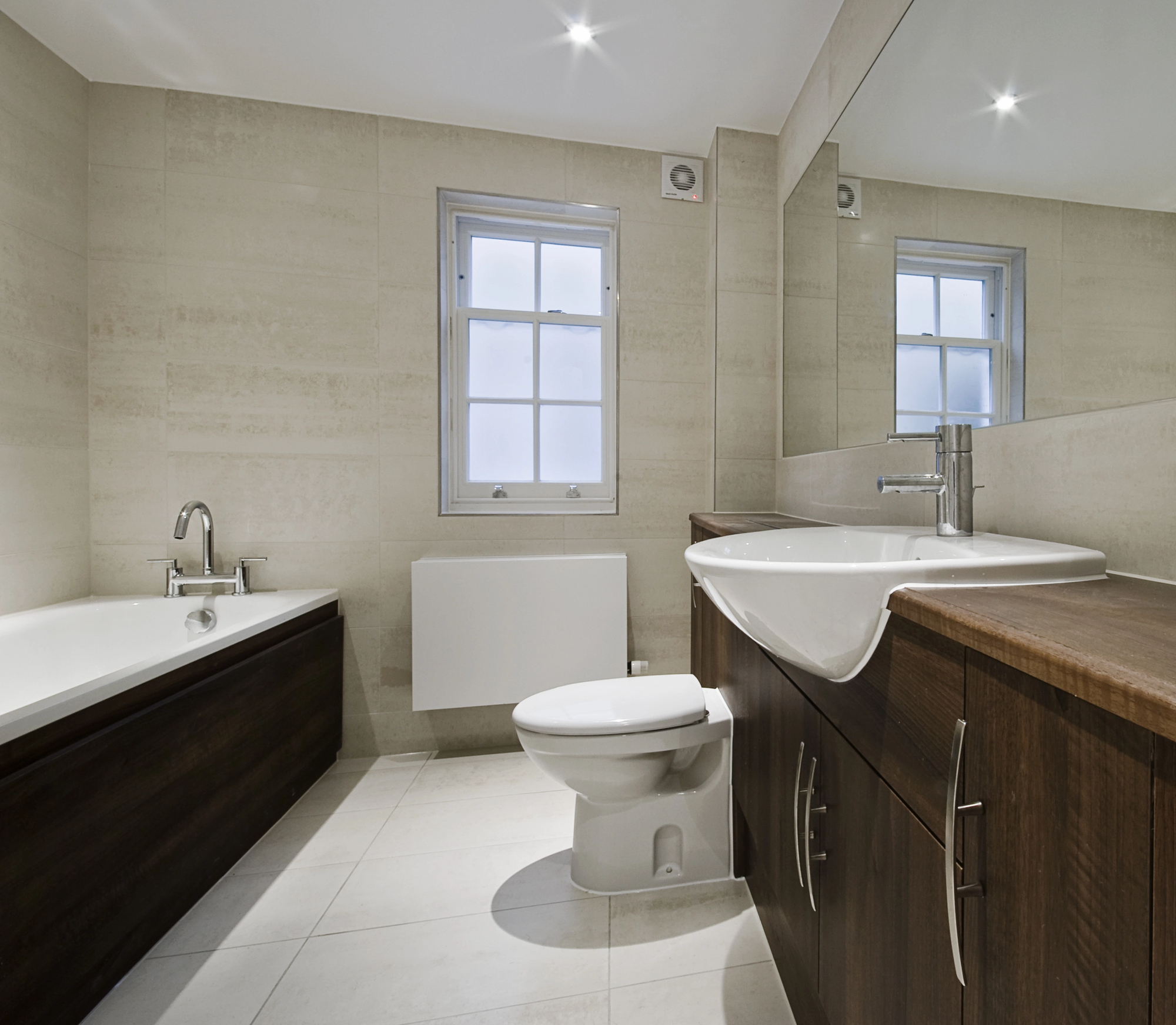 Modern bathroom with a large mirror, white sink set in dark wood cabinet, toilet, bathtub with matching dark wood panel, tiled floor and walls, and a window with frosted glass—ideal inspiration for your next bathroom renovation.