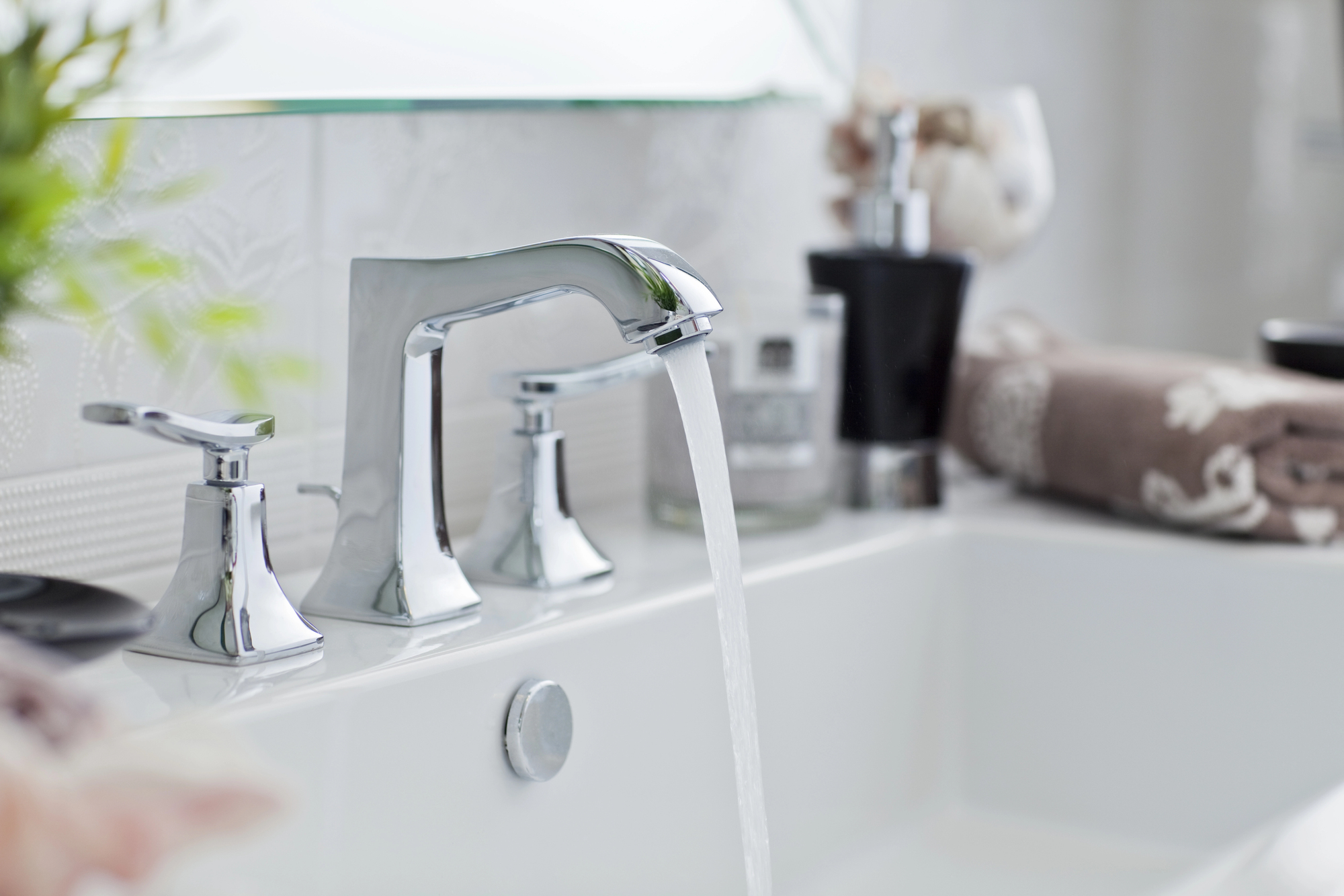 Close-up of a modern bathroom sink with chrome faucet and handles, water running into the white basin—a perfect detail shot for a BATHROOM RENOVATION or those seeking inspiration for BESPOKE BATHROOMS. Towels and decor soften the blurred background.