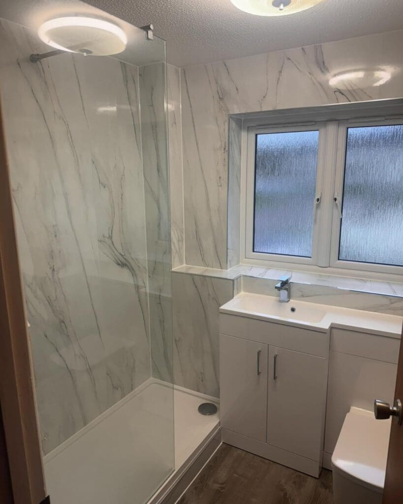 Modern bathroom with marble-patterned walls, a glass shower enclosure, and a white vanity with sink—perfect inspiration for a BATHROOM SHOWROOM. The wood-like floor and ceiling lights complete this bright, inviting space.