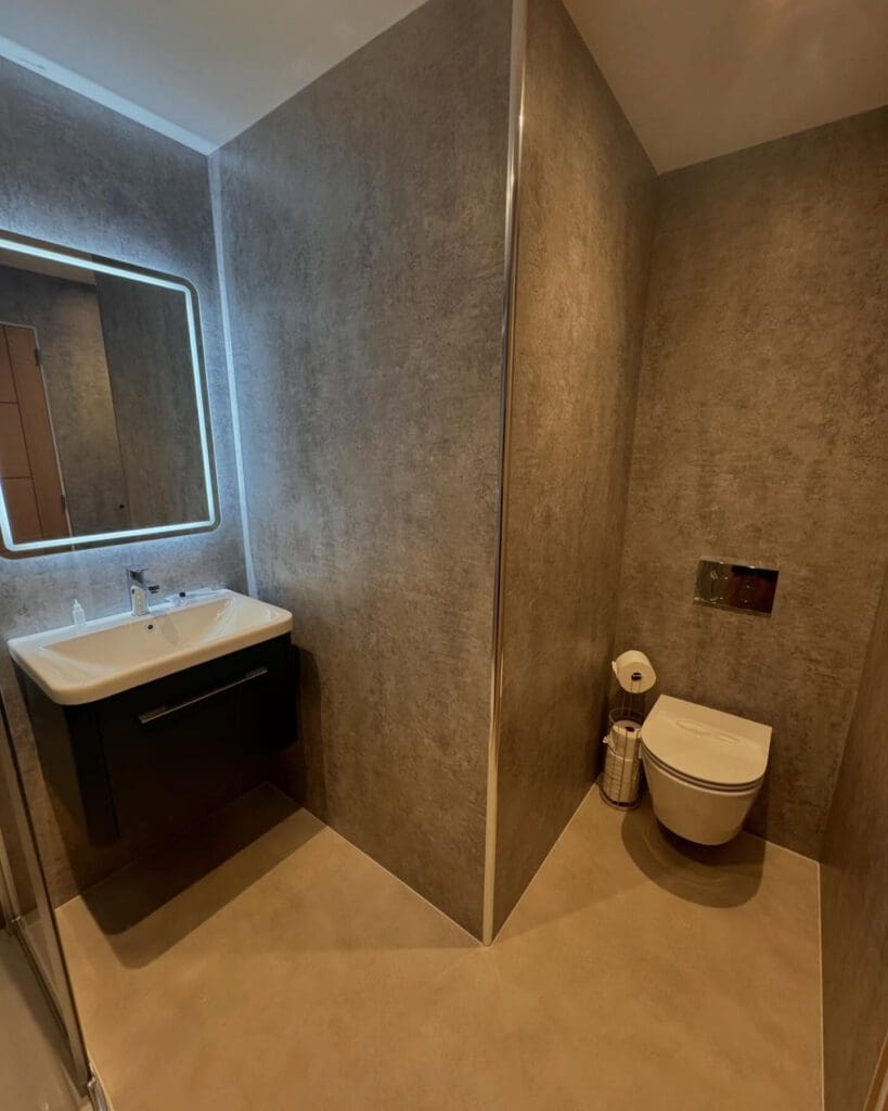 Modern bathroom with brown textured walls, perfect for a BATHROOM RENOVATION, featuring a wall-mounted sink with an illuminated mirror on the left and a wall-mounted toilet on the right, separated by a partition.