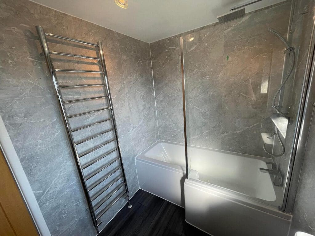 Modern bathroom with gray marble walls, a white bathtub with a glass shower screen, an overhead shower, a chrome heated towel rail, and dark flooring—perfect inspiration for your next bathroom renovation.