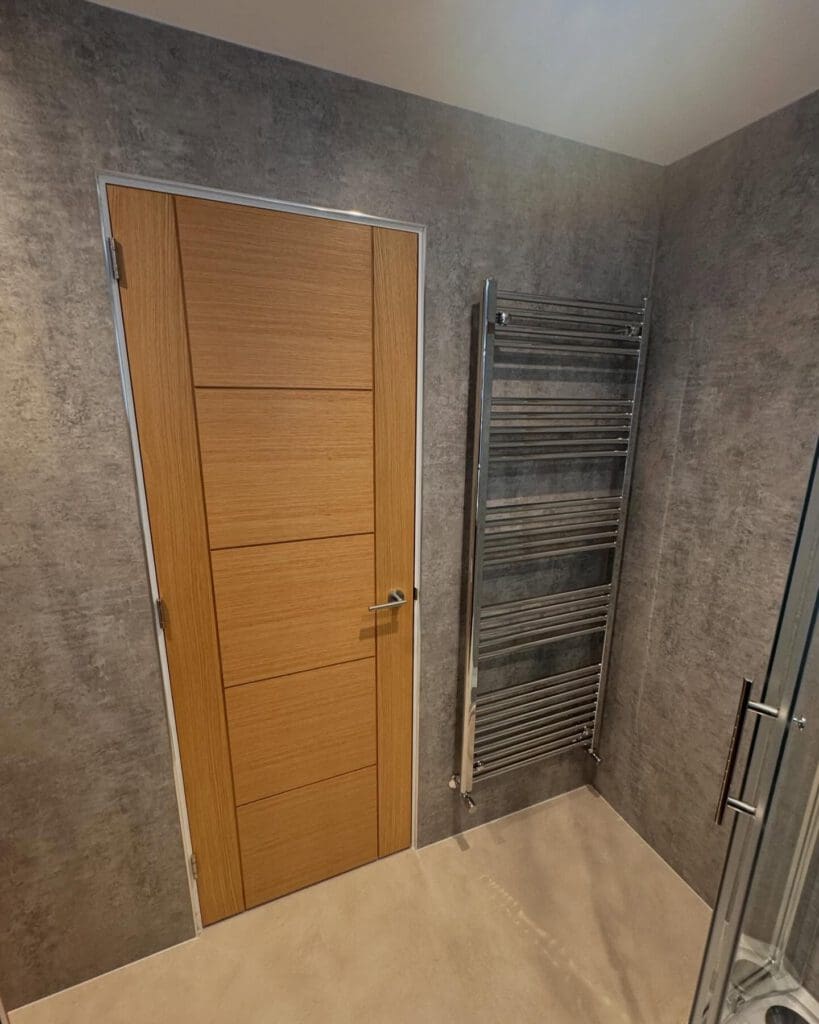 A modern bathroom with gray textured walls, a wooden door with horizontal panels, a chrome heated towel rail, and part of a glass shower enclosure hints at the stylish options available in a BATHROOM SHOWROOM for bespoke bathrooms.