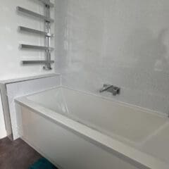 Modern bathroom with a white rectangular bathtub, silver wall-mounted faucet, and vertical heated towel rail on a speckled light gray wall. Discover BESPOKE BATHROOMS that inspire in our bathroom showroom; blue-green mat adds a pop of color to the dark floor.