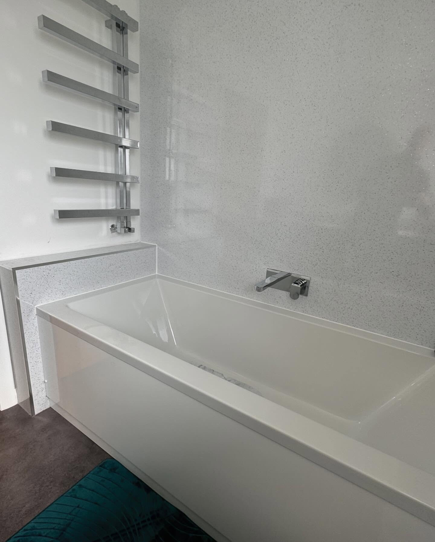 Modern bathroom with a white rectangular bathtub, silver wall-mounted faucet, and vertical heated towel rail on a speckled light gray wall. Discover BESPOKE BATHROOMS that inspire in our bathroom showroom; blue-green mat adds a pop of color to the dark floor.