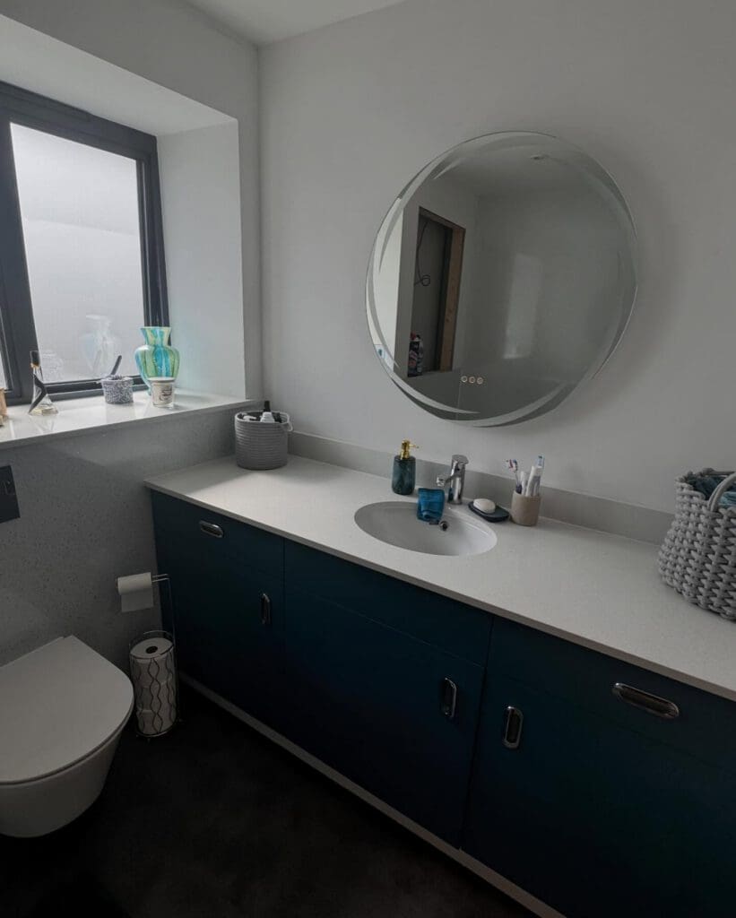 Modern bathroom with a round mirror above a white countertop, blue cabinets, and sleek sink. Discover inspiration for bespoke bathrooms with elegant toiletries, glass vases by the window, and a woven basket in this stunning bathroom showroom setup.