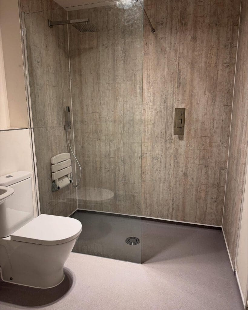 A modern accessible bathroom with a walk-in shower, wall-mounted shower seat, handheld showerhead, glass partition, and toilet. The walls have a wood-like textured finish—perfect inspiration for bespoke bathrooms or your next shower installation.