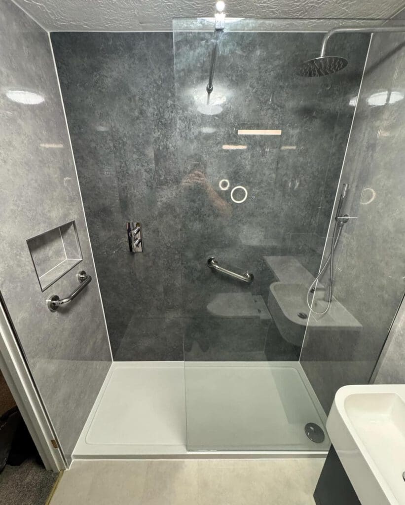 Modern walk-in shower with gray marble walls, a glass partition, rainfall and handheld showerheads, built-in shelf, grab bars, and a white shower base perfect for BESPOKE BATHROOMS in any contemporary bathroom installation.