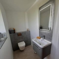 Modern bathroom showroom featuring a wall-mounted toilet, gray vanity with sink, rectangular backlit mirror, and neatly arranged towels on a gray ledge. Perfect inspiration for bespoke bathrooms and professional bathroom installation.