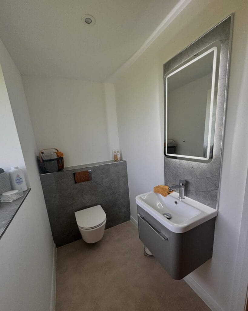 Modern bathroom showroom featuring a wall-mounted toilet, gray vanity with sink, rectangular backlit mirror, and neatly arranged towels on a gray ledge. Perfect inspiration for bespoke bathrooms and professional bathroom installation.