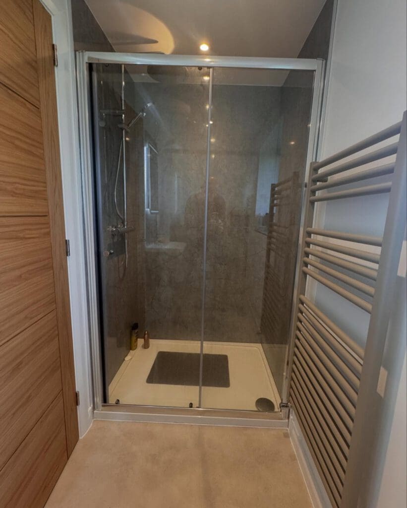 Modern bathroom renovation with a glass-enclosed shower, grey tiled walls, a wall-mounted towel radiator on the right, and a wooden door to the left. Bottles are visible in the shower corner.