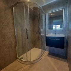 Modern bathroom with a curved glass shower enclosure, wall-mounted dark vanity and sink, illuminated mirror, and minimalist gray walls and flooring. Perfect inspiration for bespoke bathrooms or your next bathroom installation. A window reflection is visible in the mirror.
