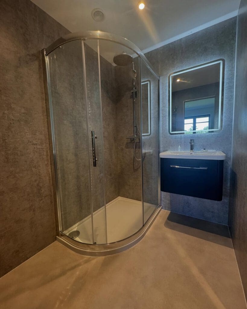 Modern bathroom with a curved glass shower enclosure, wall-mounted dark vanity and sink, illuminated mirror, and minimalist gray walls and flooring. Perfect inspiration for bespoke bathrooms or your next bathroom installation. A window reflection is visible in the mirror.