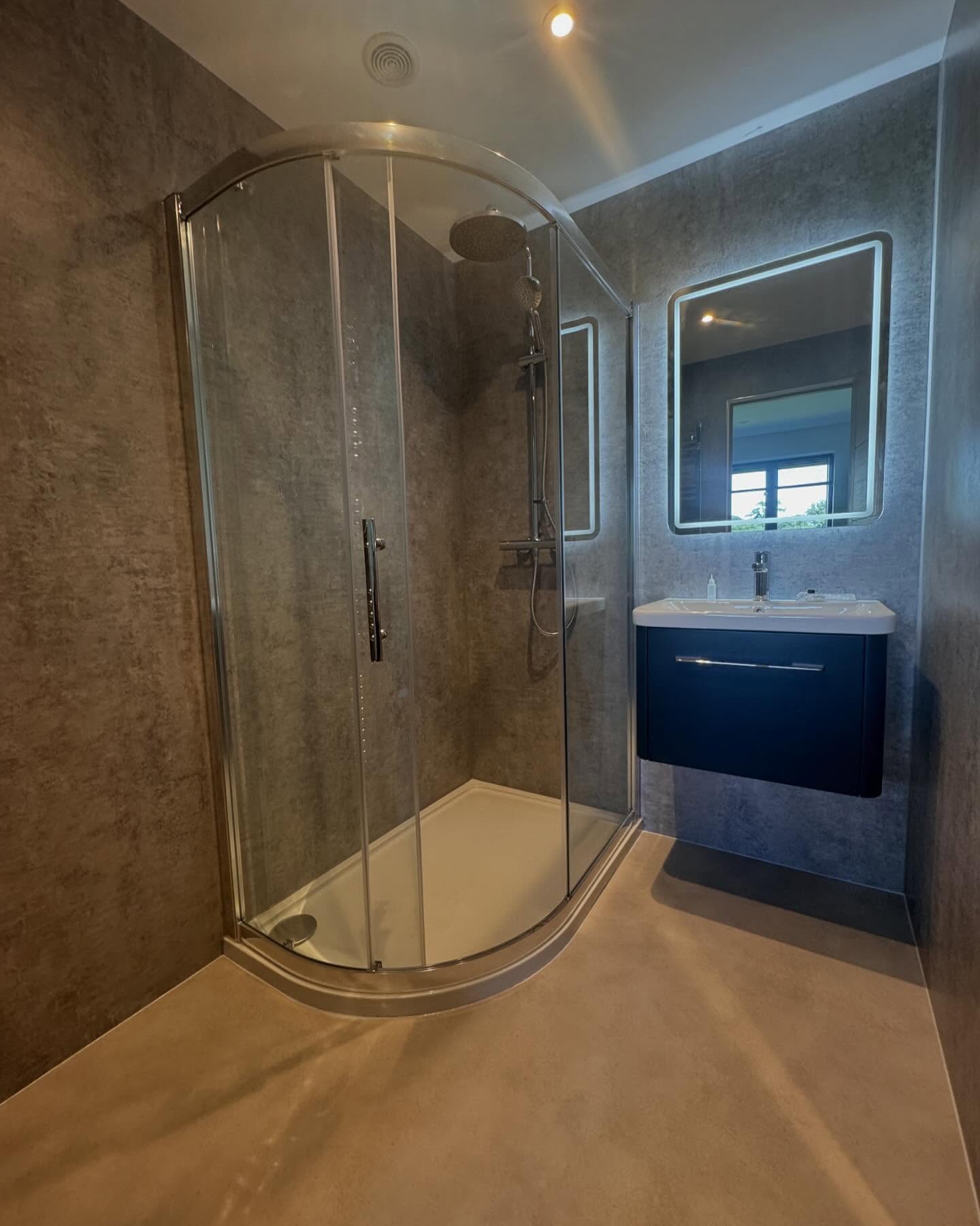 Modern bathroom with a curved glass shower enclosure, wall-mounted dark vanity and sink, illuminated mirror, and minimalist gray walls and flooring. Perfect inspiration for bespoke bathrooms or your next bathroom installation. A window reflection is visible in the mirror.